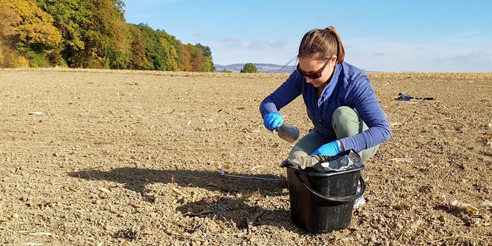 A woman wearing blue gloves and sunglasses is kneeling on a dry, plowed field while collecting soil samples in a plastic container. A black bucket is beside her. Trees with autumn foliage and a clear blue sky are visible in the background. A woman wearing blue gloves and sunglasses is kneeling on a dry, plowed field while collecting soil samples in a plastic container. A black bucket is beside her. Trees with autumn foliage and a clear blue sky are visible in the background.
