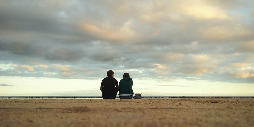  a man and a woman sitting at a beach, the endless sky above them