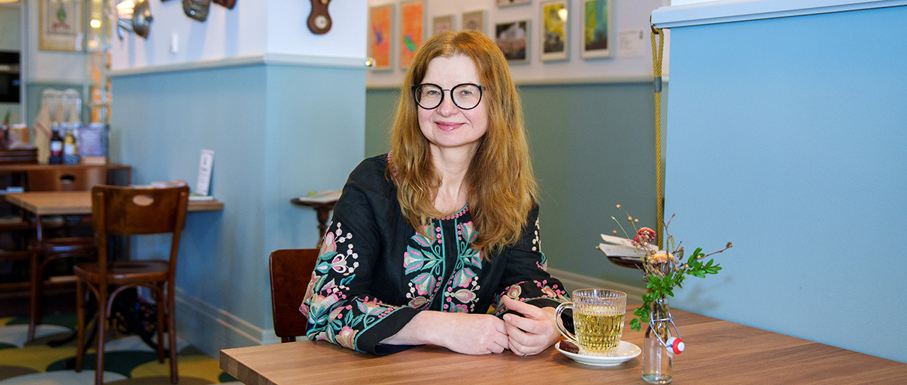 Frau mit langen roten Haaren und Brille sitzt an einem Holztisch in einem Café, vor sich eine Tasse Tee und eine kleine Vase mit Blumen; im Hintergrund hellblaue Wände und gerahmte Bilder.