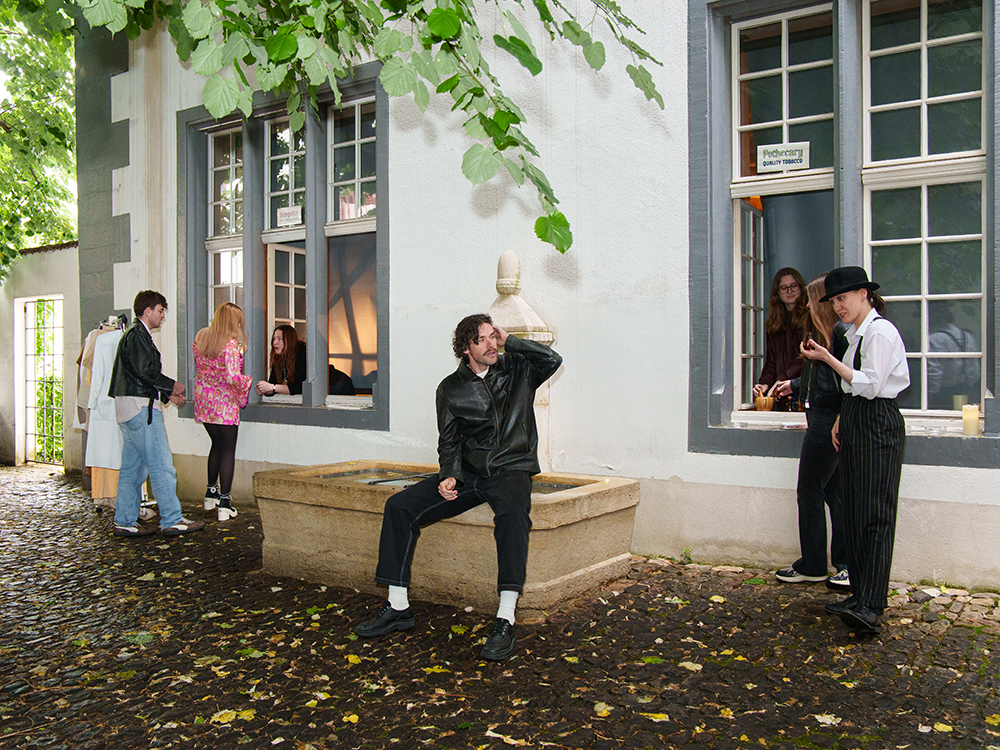 Studierende führen im Innenhof eine Szene aus einem Renaissance-Drama auf; einige stehen an Fenstern, andere befinden sich vor dem Gebäude und an einem Brunnen. Studierende führen im Innenhof eine Szene aus einem Renaissance-Drama auf; einige stehen an Fenstern, andere befinden sich vor dem Gebäude und an einem Brunnen.