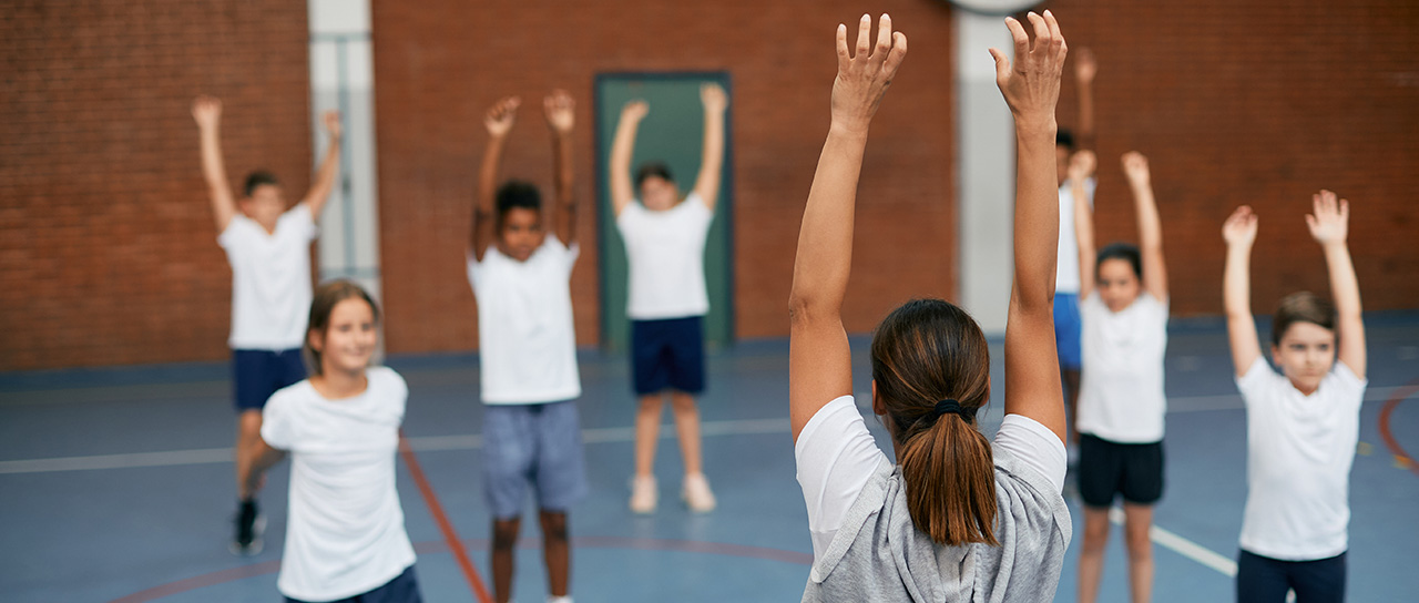 A teacher stands in a gym with their back to the camera in front of a school class; everyone is raising their hands in the air.