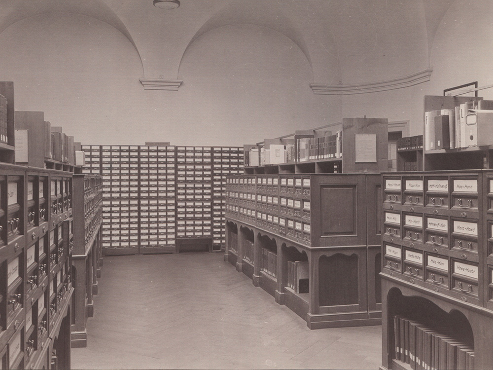 Historical black-and-white photograph of an archive or library room with tall cabinets, numerous labeled card-index drawers, and bookshelves beneath a vaulted ceiling. Historical black-and-white photograph of an archive or library room with tall cabinets, numerous labeled card-index drawers, and bookshelves beneath a vaulted ceiling.