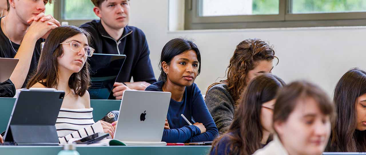 Studierende sitzen in einem Hörsaal und hören aufmerksam einer Vorlesung zu. Mehrere Personen arbeiten mit Laptops oder Tablets und machen sich Notizen.