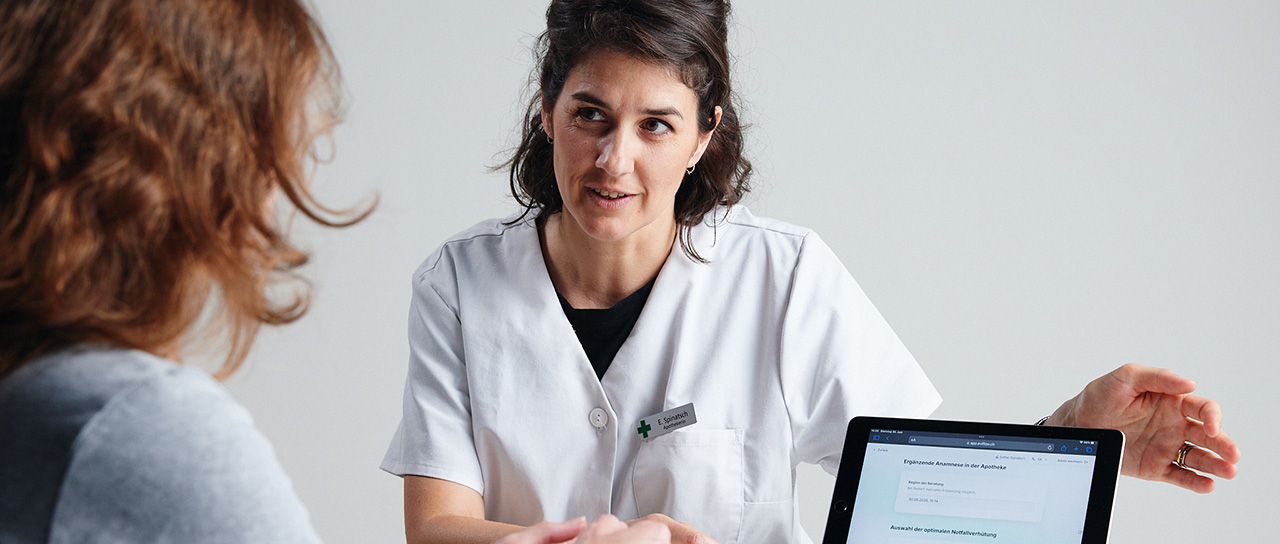 Eine Apothekerin im weissen Kittel berät eine Frau und zeigt ihr Informationen auf einem Tablet.