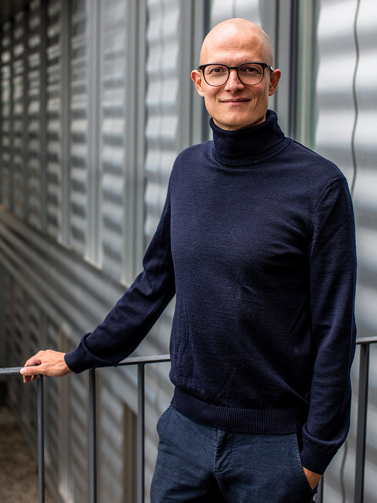 Mann mit Brille und rasiertem Kopf, der einen dunkelblauen Rollkragenpullover und eine dunkle Hose trägt, steht im Freien vor einer modernen, geriffelten Metallwand. Mann mit Brille und rasiertem Kopf, der einen dunkelblauen Rollkragenpullover und eine dunkle Hose trägt, steht im Freien vor einer modernen, geriffelten Metallwand.