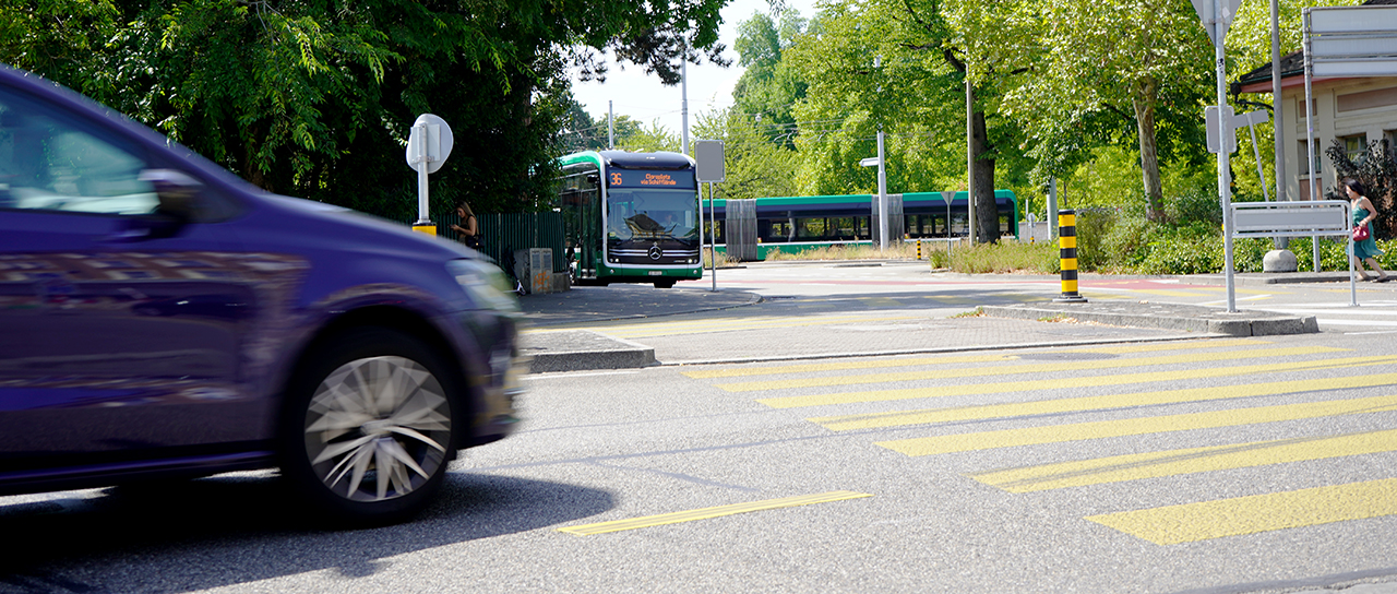 A car drives across a pedestrian crossing, with a bus and a tram visible at a stop in the background.