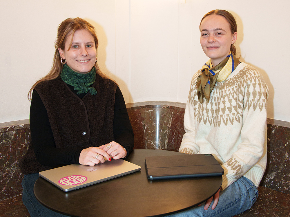 Zwei Studentinnen sitzen am Tisch und lächeln zur Kamera. Zwei Studentinnen sitzen am Tisch und lächeln zur Kamera.