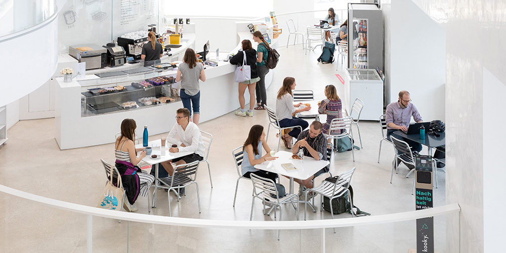 People are sitting at tables in the Biozentrum cafeteria, eating, drinking and talking. In the background, some people stand at the serving counter. 