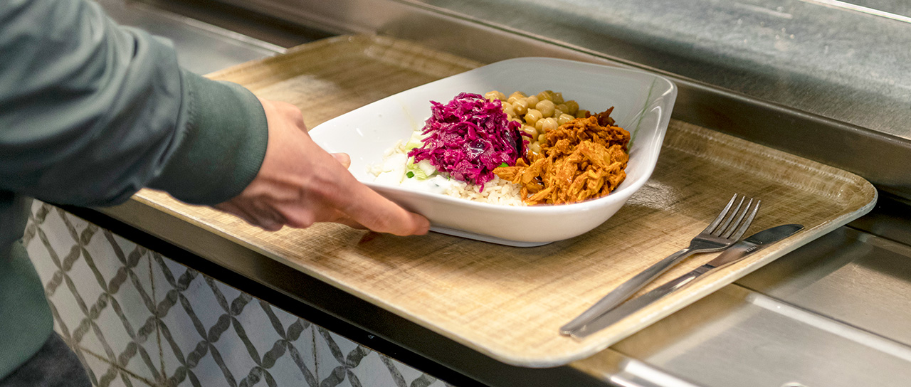 Person stellt ein Tablett mit einer weißen Schale mit Essen, darunter Rotkraut, Kichererbsen, geraspelte Karotten oder Fleisch und Reis, auf eine Cafeteriatheke. Gabel und Messer liegen auf dem Tablett.