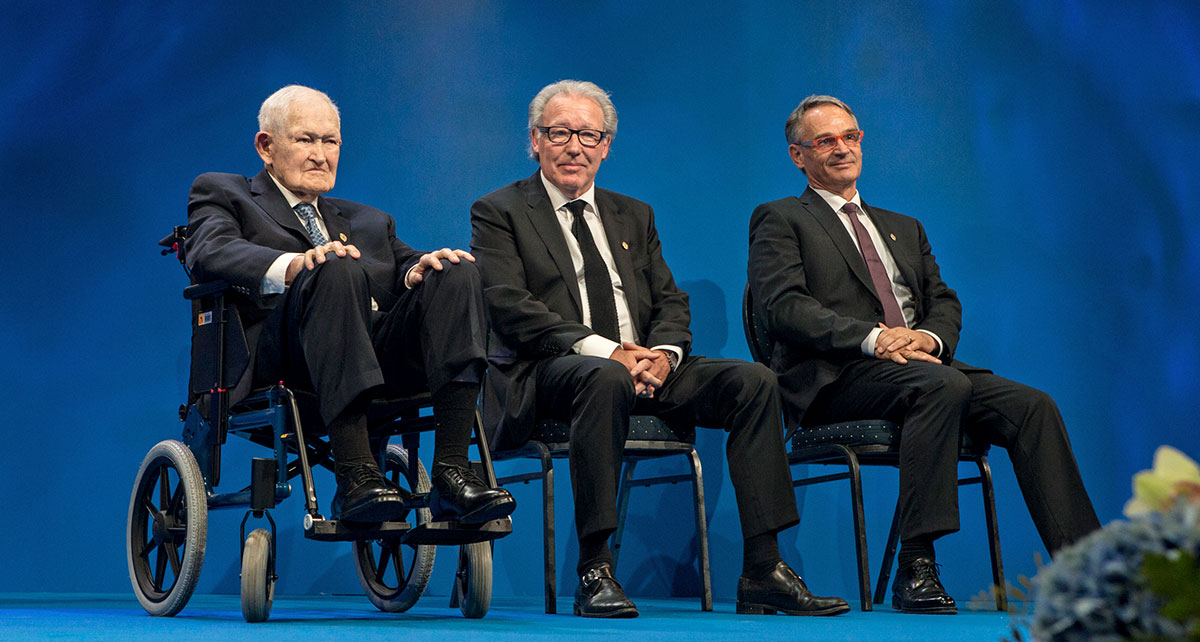 Drei Männer in Anzügen sitzen auf einer Bühne vor blauem Hintergrund; der Mann links sitzt in einem Rollstuhl, die beiden anderen auf Stühlen daneben. Drei Männer in Anzügen sitzen auf einer Bühne vor blauem Hintergrund; der Mann links sitzt in einem Rollstuhl, die beiden anderen auf Stühlen daneben.