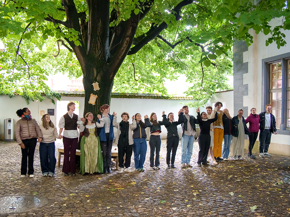 Eine Gruppe von Studierenden steht händehaltend in einem Innenhof unter einem grossen Baum und verbeugt sich nach einer Theateraufführung. Eine Gruppe von Studierenden steht händehaltend in einem Innenhof unter einem grossen Baum und verbeugt sich nach einer Theateraufführung.
