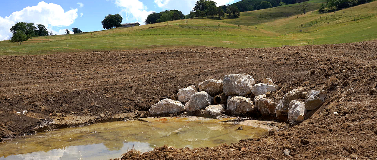 Blick auf ein trockenes Feld mit einem kleinen Rückhaltebecken, in dem sich Wasser sammelt. Steine befestigen den Zufluss am Hang.