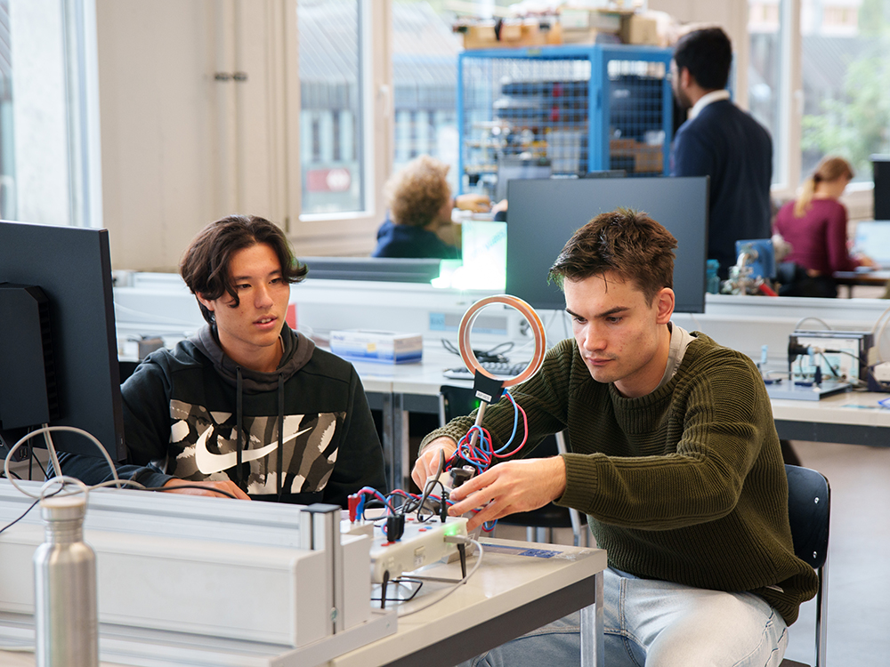 Zwei junge Männer arbeiten an einem Elektronik-Experiment mit einer Spule und verschiedenen Kabeln an einem Labortisch. Zwei junge Männer arbeiten an einem Elektronik-Experiment mit einer Spule und verschiedenen Kabeln an einem Labortisch.