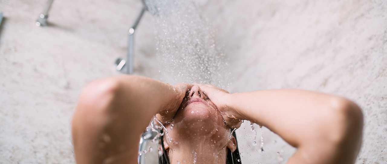 Eine Person steht unter einer Dusche, legt den Kopf in den Nacken und hält die Hände vor das Gesicht, während Wasser auf sie herabprasselt.