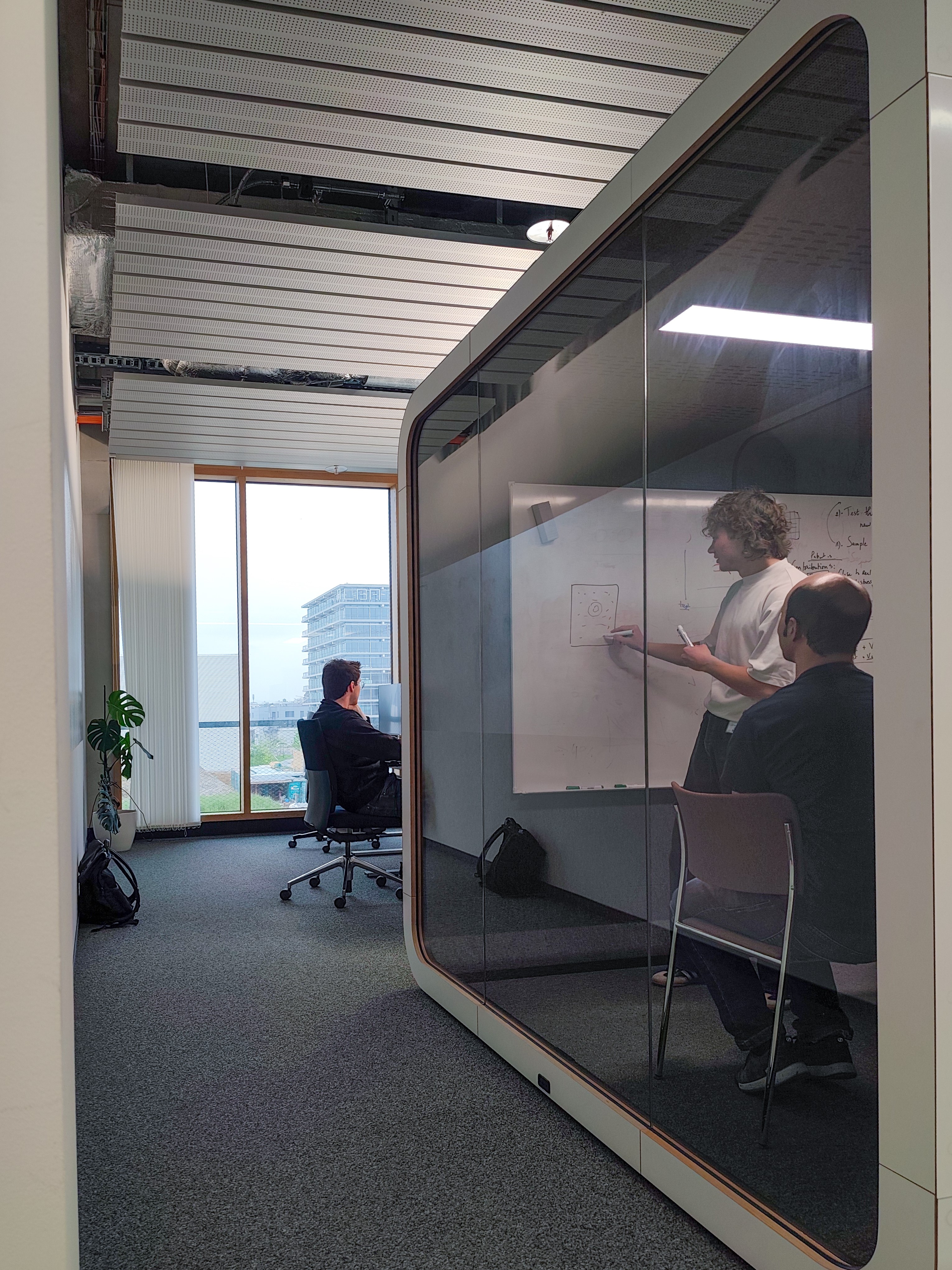 The photo shows the view of an office, with a glass cube on the right-hand side containing a whiteboard and a table with chairs. A person is drawing something on the whiteboard, someone is sitting on a chair in front of her and listening. At the end of the corridor you can see an open office space with a person sitting on a chair and looking at a monitor. The window at the end of the corridor is room-high and bright. The photo shows the view of an office, with a glass cube on the right-hand side containing a whiteboard and a table with chairs. A person is drawing something on the whiteboard, someone is sitting on a chair in front of her and listening. At the end of the corridor you can see an open office space with a person sitting on a chair and looking at a monitor. The window at the end of the corridor is room-high and bright.