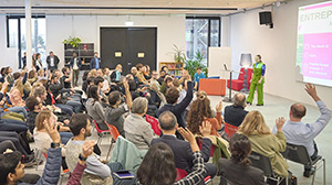 Many people sit in a room and look at a stage where a woman presents something