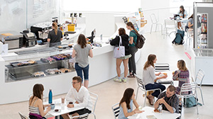 People are sitting at tables in the Biozentrum cafeteria, eating, drinking and talking. In the background, some people stand at the serving counter.