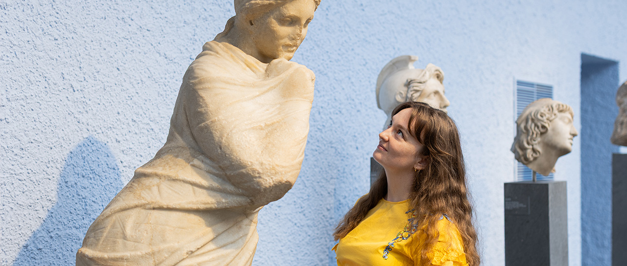 Woman in a yellow top looks at an ancient marble statue in a museum, with additional sculpted heads displayed in the background.