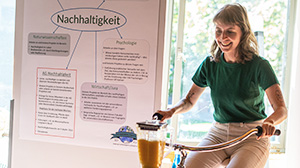 Photography of a woman sitting on a bicycle pedaling to power a blender to make smoothies. In the background is a poster introducing the AG Nachhaltigkeit