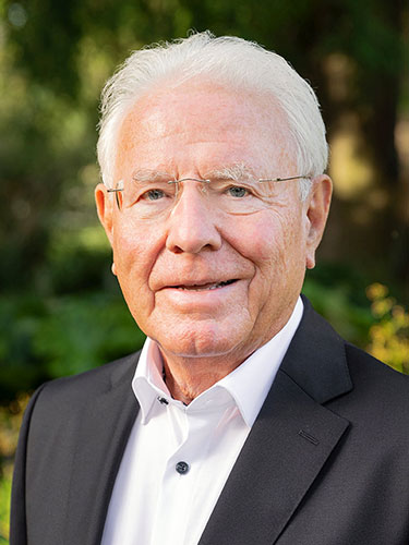 Älterer Mann mit weißen Haaren und Brille, trägt ein schwarzes Sakko und ein weißes Hemd, steht im Freien vor grünem Hintergrund. Älterer Mann mit weißen Haaren und Brille, trägt ein schwarzes Sakko und ein weißes Hemd, steht im Freien vor grünem Hintergrund.