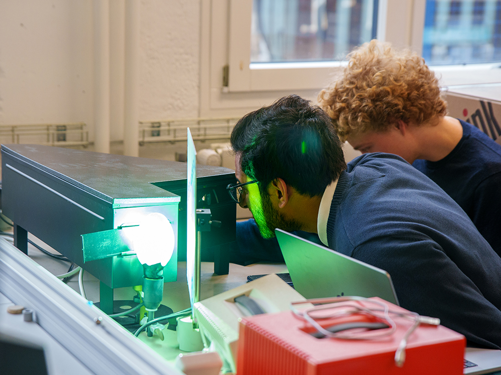 Zwei Studierende untersuchen einen optischen Versuchsaufbau mit einer grünen Lichtquelle und einem Schirm. Zwei Studierende untersuchen einen optischen Versuchsaufbau mit einer grünen Lichtquelle und einem Schirm.