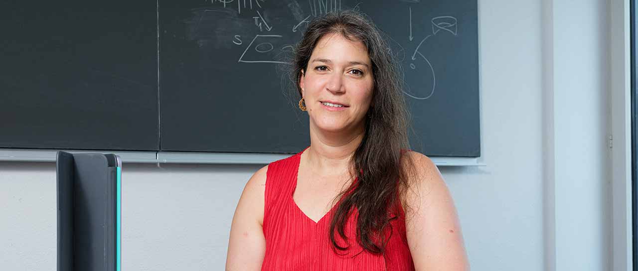 Porträt einer Frau in einem roten ärmellosen Oberteil vor einer Tafel mit mathematischen Skizzen und Formeln.