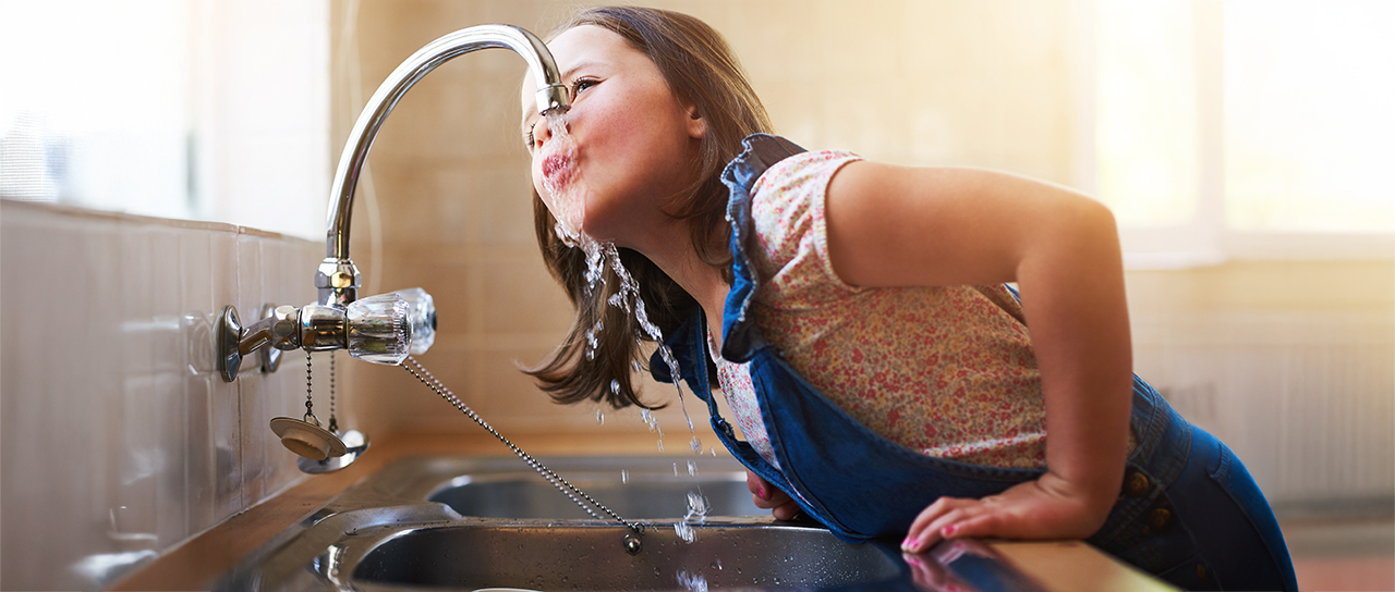 Child leaning over a sink, drinking water directly from a running tap.