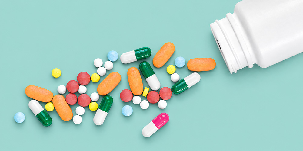 Colorful tablets and capsules are scattered next to a tipped-over white pill bottle against a mint-green background. Colorful tablets and capsules are scattered next to a tipped-over white pill bottle against a mint-green background.