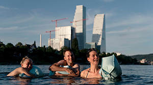 Three people are swimming in the river Rhine. In the background, modern buildings can be seen against a blue sky.