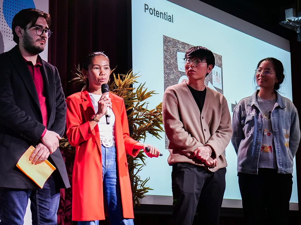 Photo of a public event organized by the Innovation Office. Four people on a podium: three men and a woman in a red coat speaking into a microphone. Photo of a public event organized by the Innovation Office. Four people on a podium: three men and a woman in a red coat speaking into a microphone.