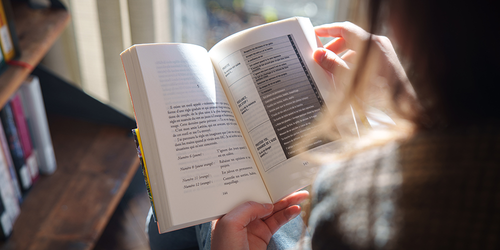 Person hält ein aufgeschlagenes Buch, von hinten fotografiert, mit sichtbar gedrucktem Text auf beiden Seiten bei natürlichem Licht.