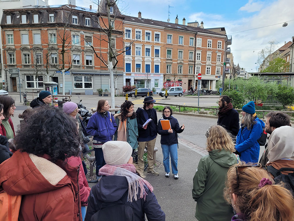 Eine Gruppe von Personen steht im Freien auf einem Stadtplatz und hört einer Person mit Brille und orangem Notizbuch zu, die etwas zu erklären scheint. Die umliegenden Gebäude haben eine Mischung aus roten Backstein- und grauen Fassaden. Eine Gruppe von Personen steht im Freien auf einem Stadtplatz und hört einer Person mit Brille und orangem Notizbuch zu, die etwas zu erklären scheint. Die umliegenden Gebäude haben eine Mischung aus roten Backstein- und grauen Fassaden.