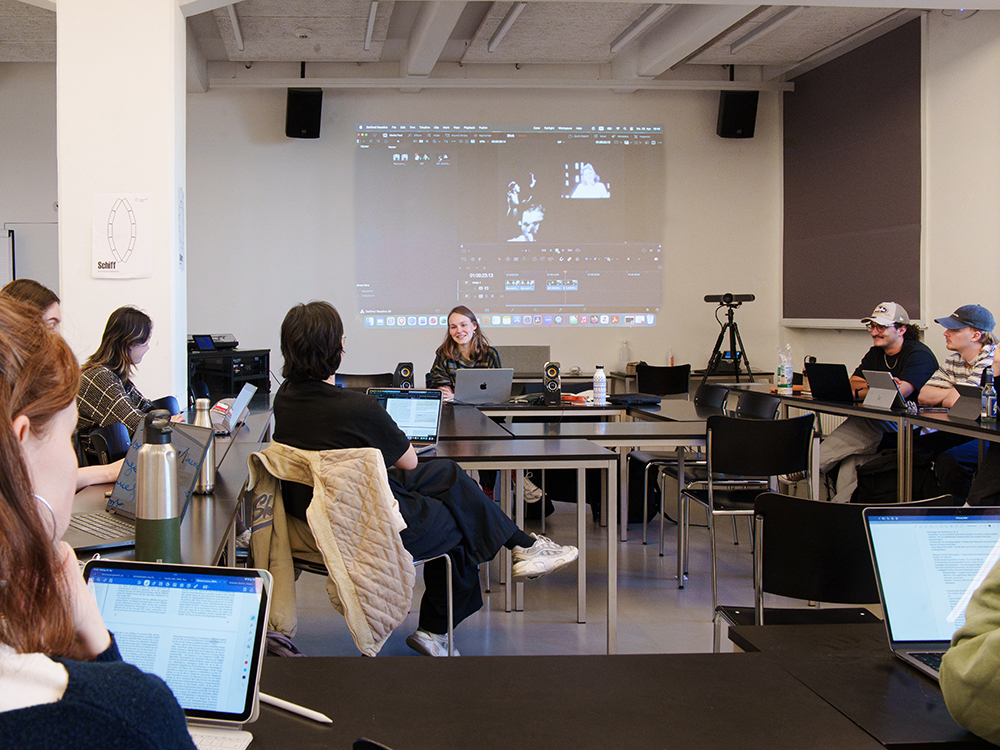 Studierende sitzen mit Laptops und Tablets in einem Seminarraum, während eine Dozentin vor einer projizierten Videobearbeitungssoftware spricht. Studierende sitzen mit Laptops und Tablets in einem Seminarraum, während eine Dozentin vor einer projizierten Videobearbeitungssoftware spricht.