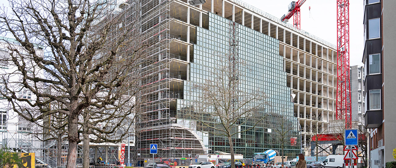 Mehrstöckiges Gebäude im Bau mit Gerüst und Kränen, an einer Straßenecke mit parkenden Autos und kahlen Bäumen.