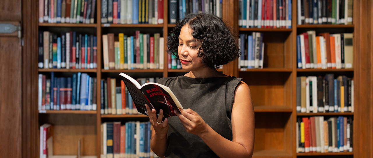 Woman with short, curly hair reading a book in front of tall bookshelves in a library.