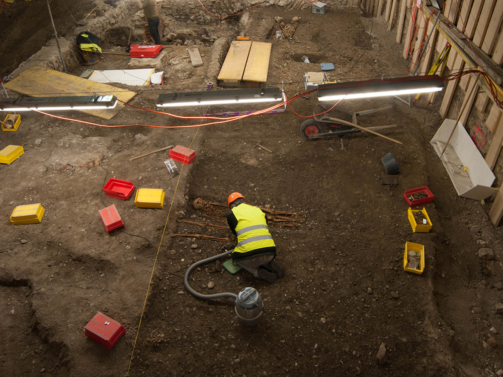 Archaeological excavation site indoors with a worker examining human remains in a trench surrounded by tools and equipment Archaeological excavation site indoors with a worker examining human remains in a trench surrounded by tools and equipment