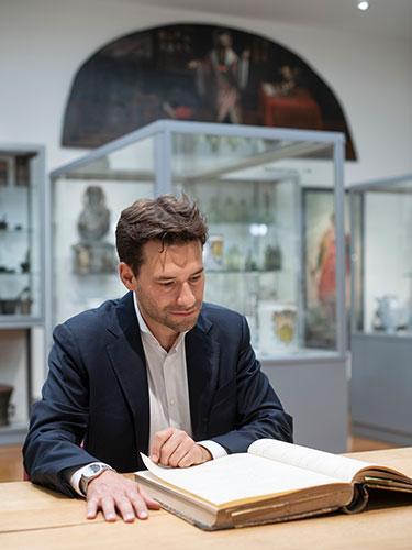 Man in a dark suit sitting at a wooden table in a museum, reading and turning pages of an old, open book. Man in a dark suit sitting at a wooden table in a museum, reading and turning pages of an old, open book.
