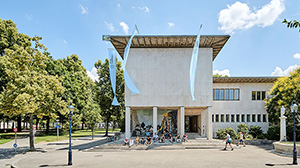 View of the entrance to the Kollegienhaus. People are sitting on the steps. Part of Petersplatz can be seen on the left. 