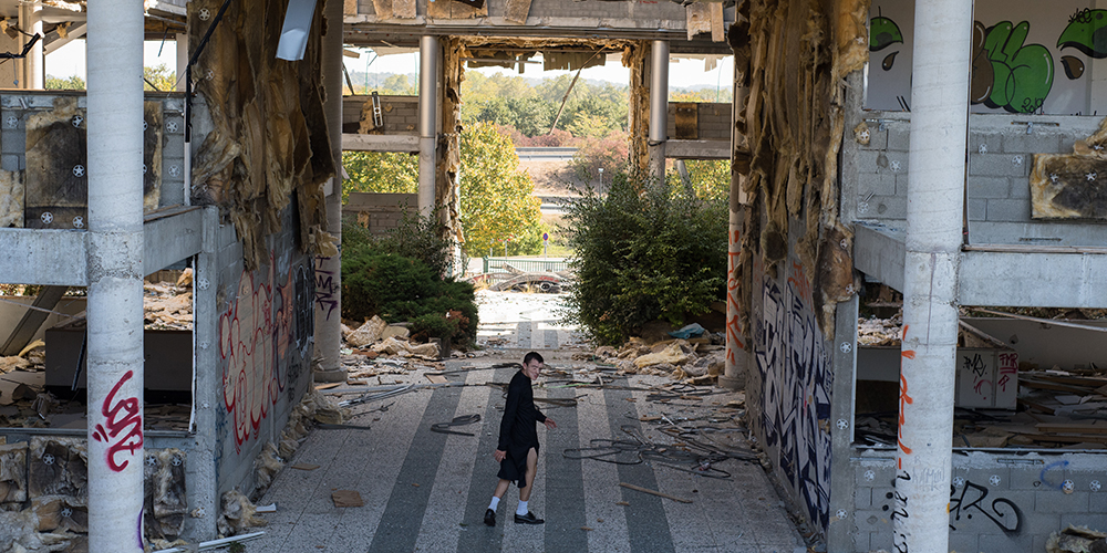 Eine Person geht durch die grosse, offene Halle eines stark beschädigten und verlassenen Gebäudes. Links und rechts sind Wände mit Graffiti, freiliegender Dämmung und Trümmern zu sehen. Im Hintergrund ist durch eine Öffnung eine Aussenanlage mit Bäumen sichtbar.