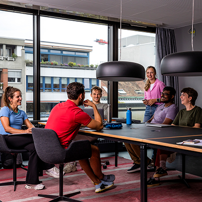 Studierende sitzen und stehen rund um einen Tisch in einem Gruppenarbeitsraum.