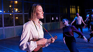 A young woman in a white blouse plays a ukulele and sings, while dancers perform in the background on a dimly lit stage.