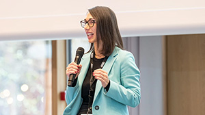 Woman in turquoise blazer talking with a microphone in hand to an convention audience 