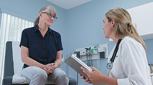 Eine Ärztin mit Klemmbrett spricht mit einer älteren Frau, die auf einer Untersuchungsliege in einer Arztpraxis sitzt.
