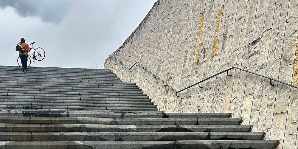 Person trägt ein Fahrrad eine breite Betontreppe hinauf neben einer hohen Steinwand mit Handläufen unter bewölktem Himmel. Person trägt ein Fahrrad eine breite Betontreppe hinauf neben einer hohen Steinwand mit Handläufen unter bewölktem Himmel.