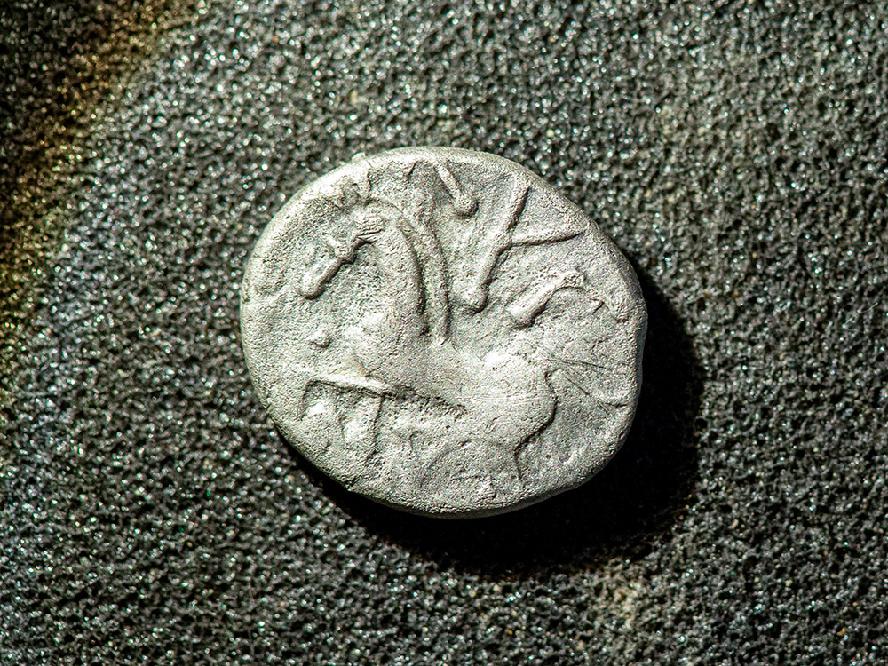 Close-up of an ancient silver coin with a worn engraving depicting a horse and rider, placed on a textured dark surface. Close-up of an ancient silver coin with a worn engraving depicting a horse and rider, placed on a textured dark surface.