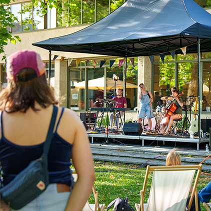 Menschen sitzen im Freien vor einer kleinen Bühne, auf der eine Band spielt.