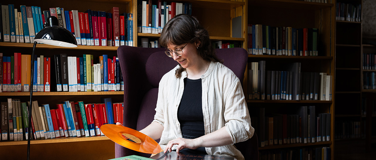 Lavinia Pflugfelder sitzt auf einem Stuhl vor einem Bücherregal und schiebt eine orangefarbene Schallplatte in die Hülle zurück. Auf dem Tisch vor ihr liegen weitere Schallplatten sowie ein dickes Buch.