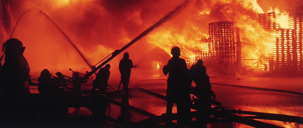 Feuerwehrleute bekämpfen nachts einen Grossbrand, während eine Lagerhalle hinter ihnen in hellen Flammen steht.