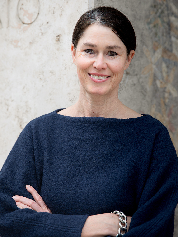 Eine Frau mit dunklen, zurückgebunden Haaren trägt einen dunkelblauen Pullover und lächelt. Sie steht vor einer großen, leicht unscharfen Mosaikwand mit floralen und natürlichen Motiven. Eine Frau mit dunklen, zurückgebunden Haaren trägt einen dunkelblauen Pullover und lächelt. Sie steht vor einer großen, leicht unscharfen Mosaikwand mit floralen und natürlichen Motiven.