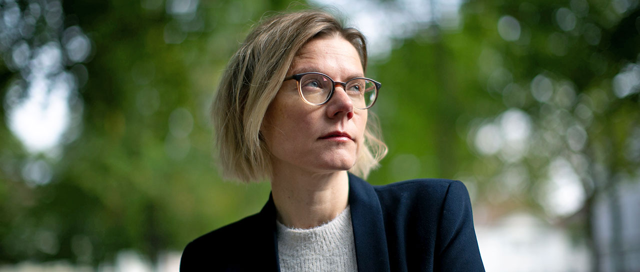 Portrait of a woman with short blonde hair and glasses, wearing a dark blazer over a light grey sweater.